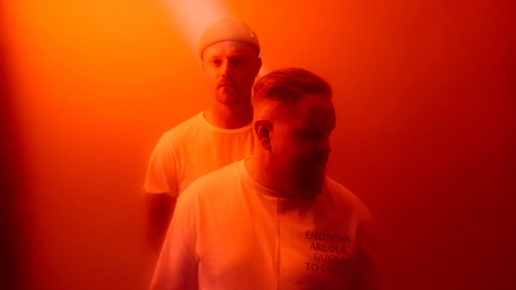 Photo of the two members of anamē wearing white shirts against a hazy orange background