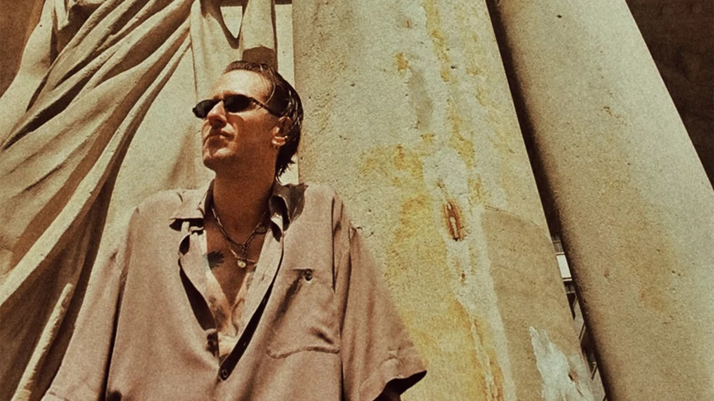 Photo of Luca Venezia sitting against a pillar wearing a beige shirt and sunglasses