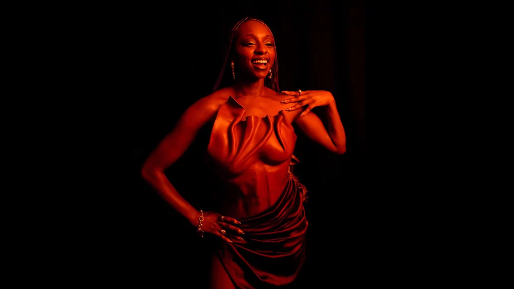 Photo of Kikelomo posing against a black background. She's smiling and wearing a brown-gold dress and is illuminated in a red light