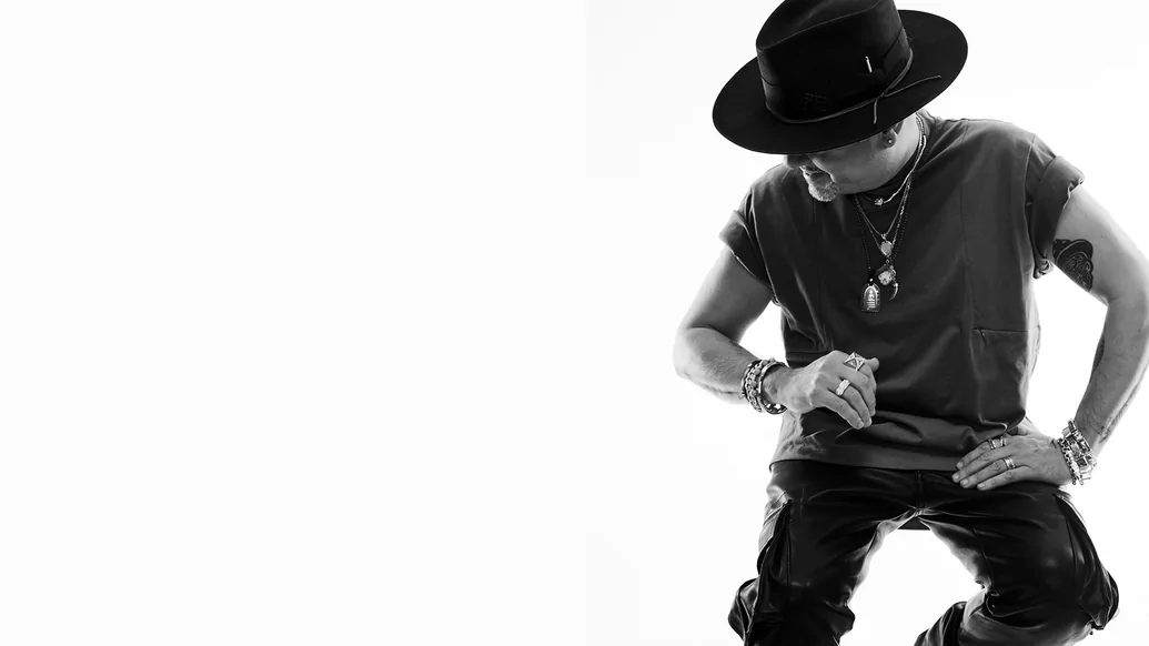 Black and white photo of Louie Vega wearing a black hat and t-shirt while sitting on a chair