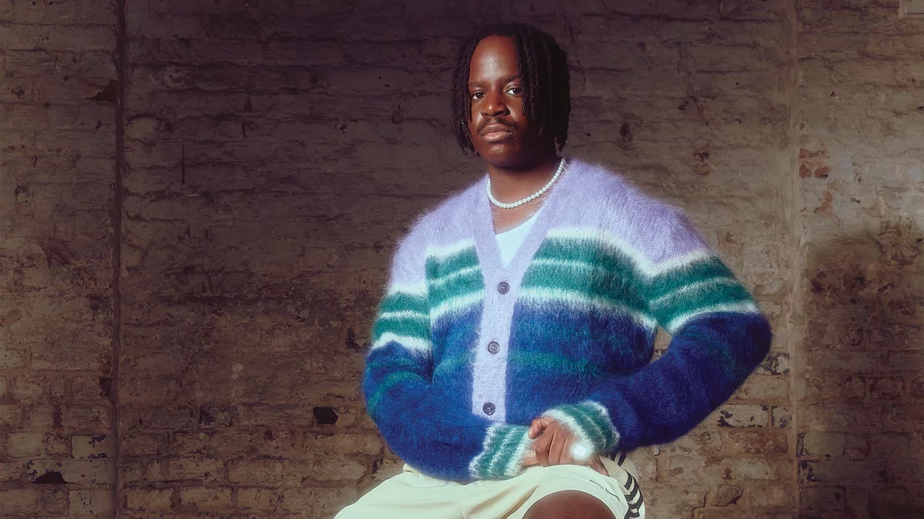 salute sitting on a stool in front of a brick wall wearing a a blue, green and lilac fuzzy sweater