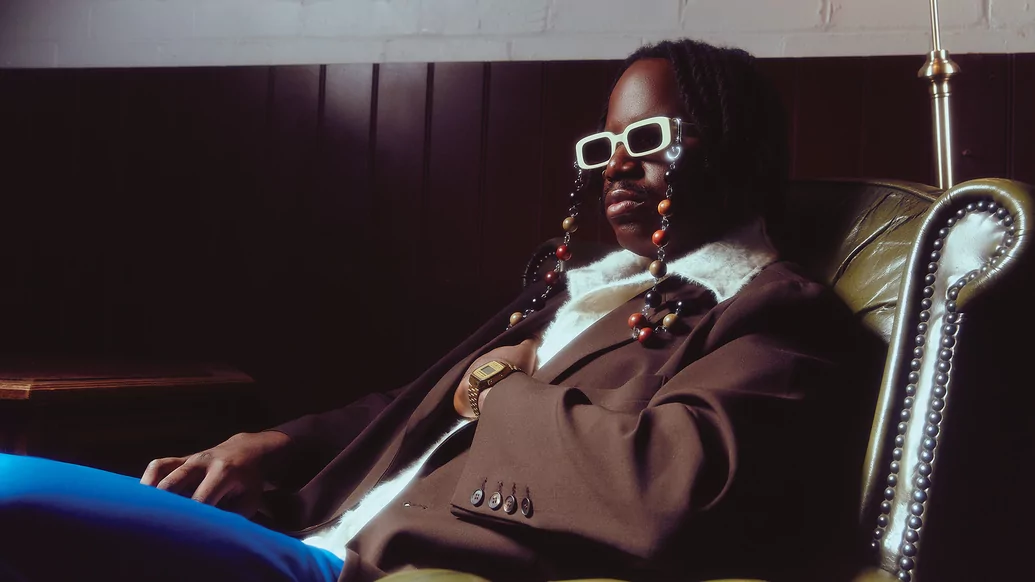 salute reclining in an antique green leather wearing white sunglasses with beads hanging from them and a black suit jacket