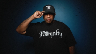 Press shot of DJ Premier in a black hat looking toward the camera in a t tshirt that says "Royalty"