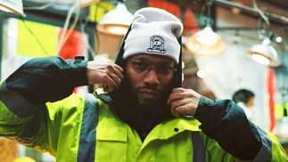 Photo of WTCHCRFT wearing a hi-vis jacket and a white beanie. He's wearing glasses and looking intently at the camera while holding his collar in his hands