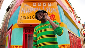 Photo of Ekiti Sound wearing a green wooly sweater with black and yellow strips. He's on the phone and wearing a bucket hat and sunglasses. He's standing in front of a corner shop painted light blue and red