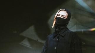 Photo of Baby T wearing a black coat and balaclava in front of a shadowy background