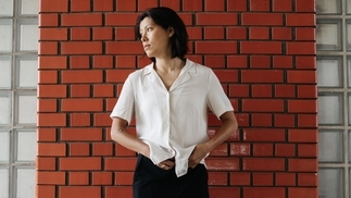 Photo of Monki wearing a white shirt and black trousers against a faux brick wall