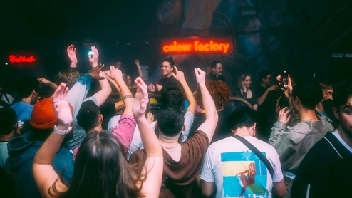 Photo of Danielle DJing taken from the dancefloor in Colour Factory. The club's sign hangs behind her and people in the crowd have their hands in the air