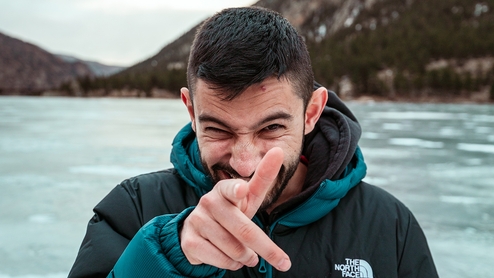 Hamdi standing in front of a lake wearing a north face jacket and pointing at the camera