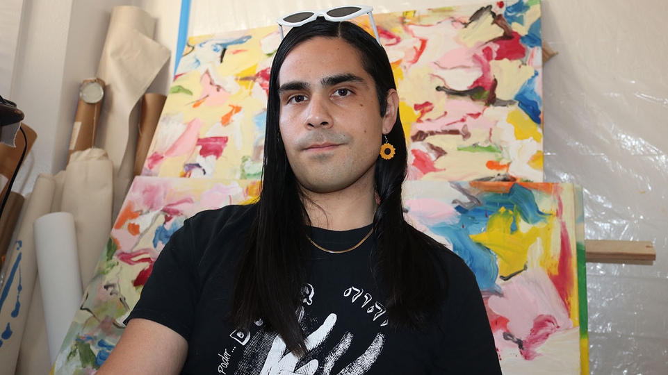 Photo of Farsight sitting in front of a colourful abstract painting. His hair is long and black and he has sunglasses resting on his head. He's wearing a dangling orange earing. 
