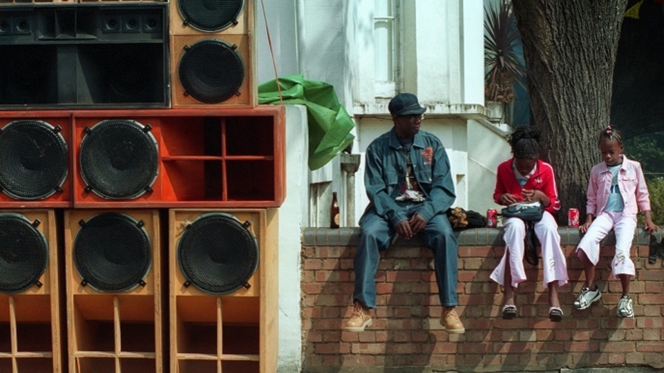 Photo of three people sitting next to wall of sound systems at Notting Hill carnival